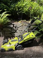 Side view of Ryobi electric lawnmower in outdoor garden setting showing full mower and attached grass bag.