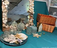 Photo showing the wooden framed mirror decorated with glued seashells, 2 woven square baskets, seashells displayed on a plate, and ceramic candle holders.