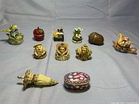 View showing 10 small ceramic and resin decorative boxes arranged on flat surface including floral, fruit shapes, and royal trio figurines.
