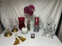 Wide view of all items showing brass candle holders, red glass vase, white ceramic vase, pewter candle holder with candle, glass rectangular vase with flowers and stones, golden tassels, and decorative floral cubes.