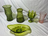 Four olive green glass pieces and one pink glass vase displayed against a white cloth background. The green pieces include textured and smooth vases and two uniquely shaped bowls. The pink vase has embossed floral and geometric designs.