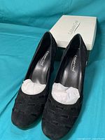 Front view of pair of Coldwater Creek black leather wedge shoes with cutout design and white stuffing inside, placed on turquoise background with original box behind.