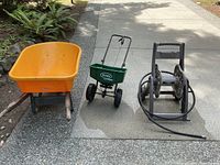 Yellow True Temper wheelbarrow, green Scotts seed spreader, and gray hose reel with attached hose lined up on pavement.