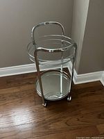Front angle view of round metal and glass bar cart with two shelves and caster rollers, showing the frame and glass surfaces clearly.