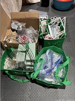 Four green bags and brown box filled with floral foam, wire frames, zip ties, stones, beads, and other crafting supplies.