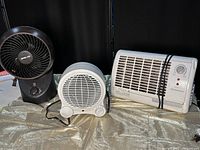 Three items on a table against a dark background: a black ORECK fan, a small white fan, and a white rectangular heater with a cord wrapped around it.