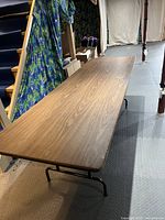 Long wooden folding table with black metal legs shown in basement environment, displaying general wear on top surface.