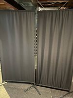 Two black privacy screen panels side by side, showing the fabric and metal frame with tripod base.
