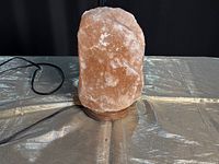 Large Himalayan salt lamp sitting on a wooden base with a visible power cord placed on a reflective silver surface under neutral lighting.