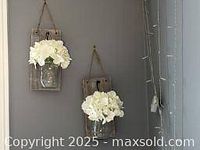Two photos showing the same wall decor setup featuring two mason jars with white faux flowers mounted on wooden plaques hanging on a wall near curtains and lights.