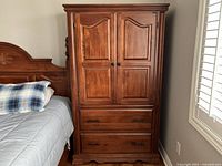 Front and side view of the wooden wardrobe showing two paneled doors on top and two drawers below. Positioned next to a bed.