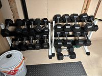 Two dumbbell racks loaded with assorted dumbbells from 8 lb to 25 lb, with bucket and mat in foreground