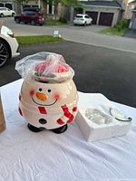 Front view of ceramic snowman candy jar with lid wrapped in plastic, showing smiling face, red scarf and buttons.