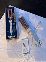 New Conair CD704 Double Ceramic Triple Barrel Hair Styling Tool with box, attached power cord, and instruction manual laid out on dark surface.