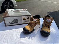 Pair of tan leather Dolphin brand safety boots with black reinforced steel toes on white surface with box in background. Size 4 indicated on box.