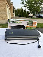 FoodSaver FM2000 vacuum sealer in front of its box, placed on a white cloth outside. Device and box visible with power cord.