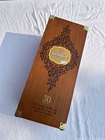 Top view photo of the Glenfiddich wooden whisky box showing intricate black floral designs and central gold Glenfiddich logo plaque, along with '30' marking and corner metal protectors.