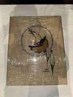 Front view of framed abstract artwork with a circular design including stylized shapes and feathers, wrapped in plastic.