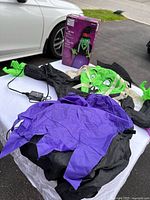 Inflatable witch decoration laid out with box, power adapter, showing green witch face, black hat, purple and black dress, and green hands.