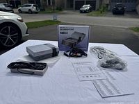 Photo of game console, wired controller, power adapter, AV cable, user manual, and box on white cloth outdoors.