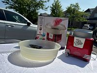 Full view of the red Nostalgia cotton candy maker with its clear plastic bowl and box outdoors on a white table.