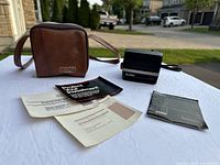 Polaroid Sun 600 LMS instant camera with leather carrying case and manuals laid out on white table outdoors.
