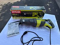 Ryobi P514 reciprocating saw with box and manual laid out on white cloth outdoors, showing front and side views with power cord visible.