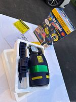 Laser light projector in black with green laser band, placed in white protective styrofoam inside the original box. Instruction manual and warning labels beside it.