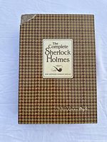 The front of the slipcase showing 'The Complete Sherlock Holmes' with a pipe icon on a brown houndstooth pattern, corner tear visible at top left of slipcase.