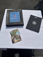 Overall view of art book, storage box, and two abstract prints laid flat on white surface outdoors.