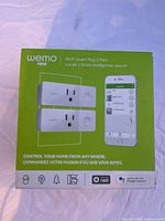 Front of product box showing two Wemo Mini Wi-Fi smart plugs and a smartphone app interface on green background.