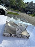 Photo of the Wild Eye Designs Brie cheese baker set in original clear plastic packaging outdoors on a table, showing white ceramic items inside and some box damage.