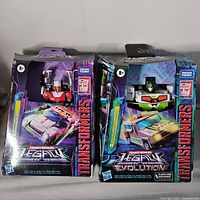 Photo of two new Transformers Legacy figures in original Takara Tomy and Hasbro packaging showing front view of boxed figures, with colorful artwork and windows.