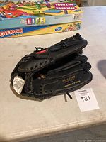 Photo of a folded black leather baseball mitt with tag, showing part of the woven basket-style pocket and printed text including model number PM1250B and 'Leather Palm'. The mitt is positioned on a table with board games in the background.