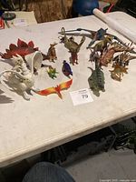 Collection of various dinosaur figurines arranged on white table including winged and spiked dinosaurs in multiple colors and sizes.