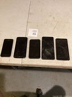 Five used phones lined up on table. Various models including iPhones. Four phones have cracked or shattered screens.