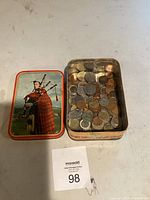 Top-down view showing open tin filled with assorted old foreign coins and lid featuring a Scottish bagpiper.