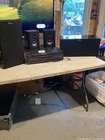 Photo showing the multi disc CD player surrounded by two bookshelf speakers and two larger speakers on a folding table in a garage or similar setting, daylight visible behind.