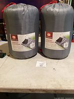 Two gray Outbound sleeping bags in compression sacks with red drawstrings on table.