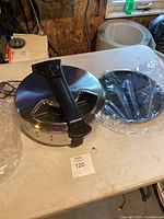 Front and side view of electric pizza cooker with glass lid and black handle on table next to round black plastic accessory.