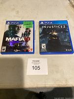 Two PS4 game cases on white surface with a lot number card 105. Left case Mafia III, right case Injustice 2.