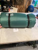 Photo of green rolled sleeping mat showing full length, secured with two black elastic straps, placed on a white table.