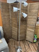 Full view of the standing lamp showing all three light fixtures with frosted glass shades and brushed metal pole and round base, placed in front of a wooden folding screen and next to a grey couch.
