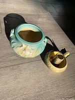 View of Roseville turquoise floral iris vase and yellow textured Ineke ceramic candleholder on floor with shadow.