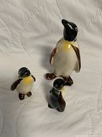 Set of three ceramic penguin figurines displayed on a white quilted surface showing size and color details