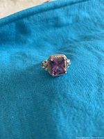 Photo of silver-colored metal ring with rectangular simulated amethyst stone, resting on blue fabric background.