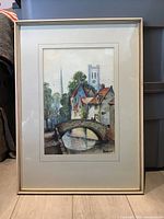 Framed watercolor painting showing an architectural scene with a stone bridge, canal, houses, and church towers in the background.