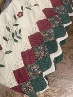 Close-up of scalloped edges showing patchwork squares with floral and patterned fabrics in burgundy and green tones, with a white scalloped edge featuring a red pattern.