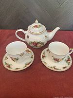 Front view of teapot with lid, two matching tea cups and saucers arranged on red fabric background with dark herringbone wall.