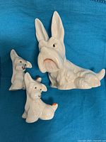 Three white Beswick porcelain dog figurines, one larger lying dog and two smaller sitting dogs, all with hand-painted ladybugs, set on a blue fabric background.